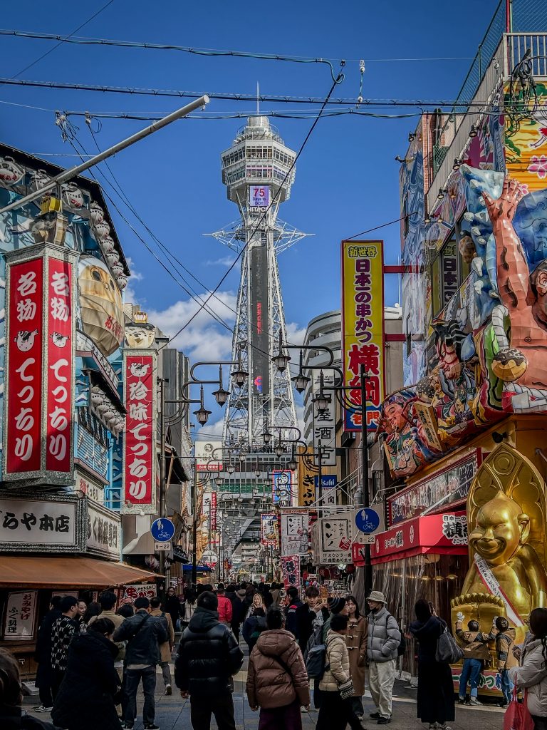 Rua movimentada em Osaka, Japão, com a icônica Torre Tsutenkaku ao fundo, cercada por letreiros vibrantes em japonês, lojas tradicionais e restaurantes. O cenário colorido inclui estátuas e murais chamativos, enquanto pedestres caminham e exploram o ambiente urbano animado.