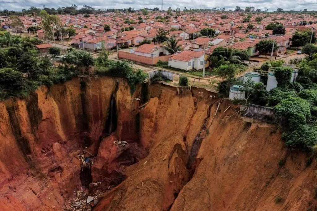 Imagem aérea mostra uma enorme cratera de erosão próxima a um bairro residencial com casas de telhado vermelho, evidenciando o risco geológico iminente para a comunidade.