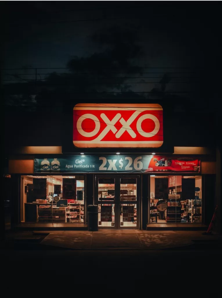 Loja de conveniência OXXO iluminada durante a noite, com destaque para o letreiro vermelho e branco e uma promoção de água mineral 2x$26 no topo da entrada.