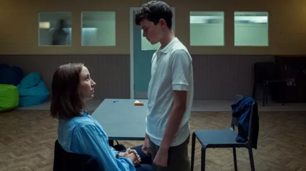 Cena da série Adolescência em que a advogada de Jamie o confronta durante uma visita ao centro de detenção. A troca de olhares intensa expõe a gravidade do momento: um adolescente acusado de homicídio, diante das consequências reais de seus atos.