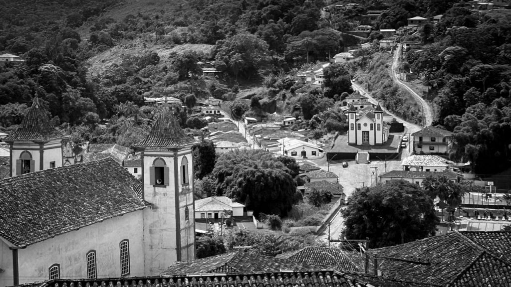Vista panorâmica da cidade histórica de Serro, em Minas Gerais, com igrejas coloniais, telhados de barro e morros ao fundo sob céu com nuvens densas.
